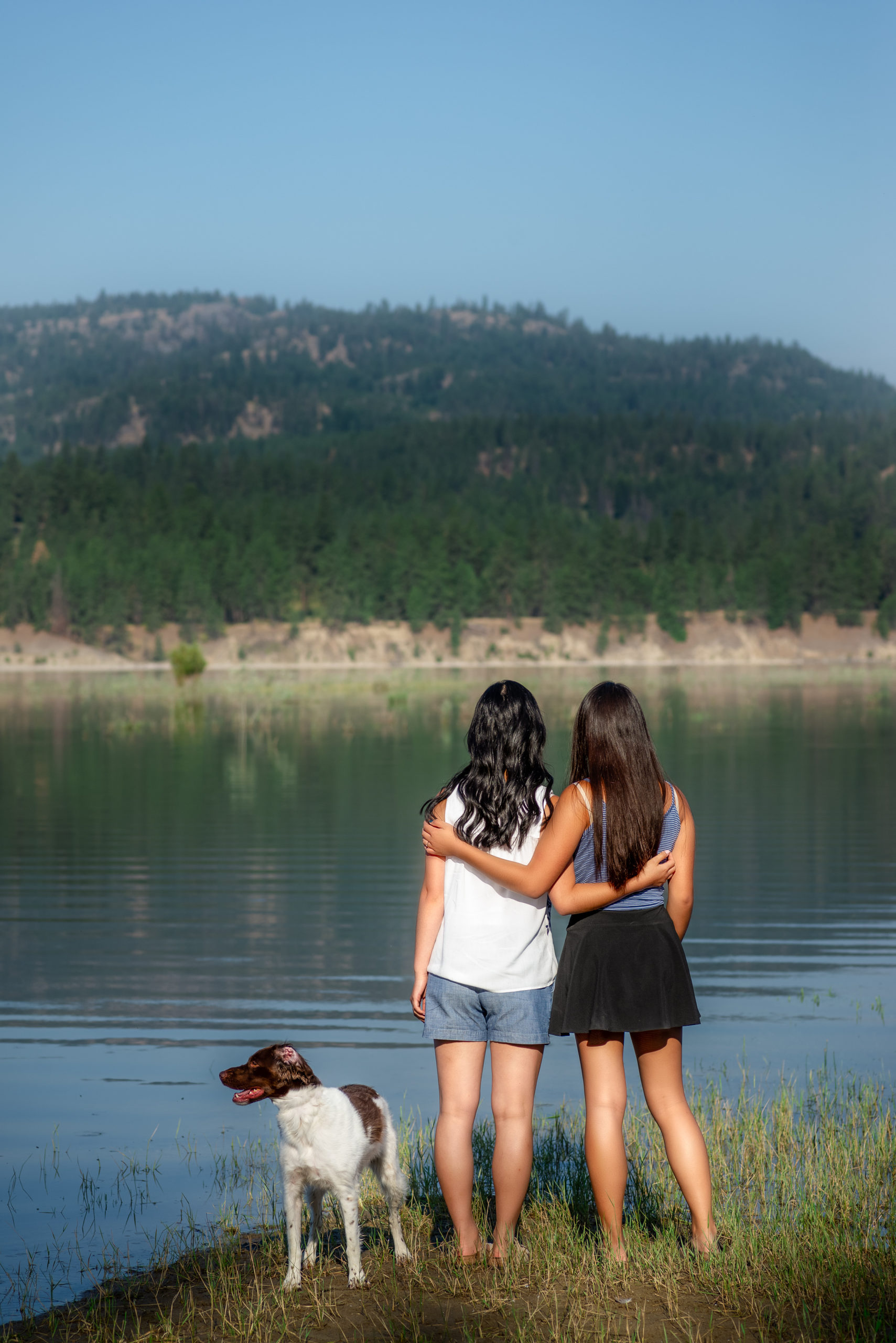 sisters hugging each other with their dog while looking at the lake