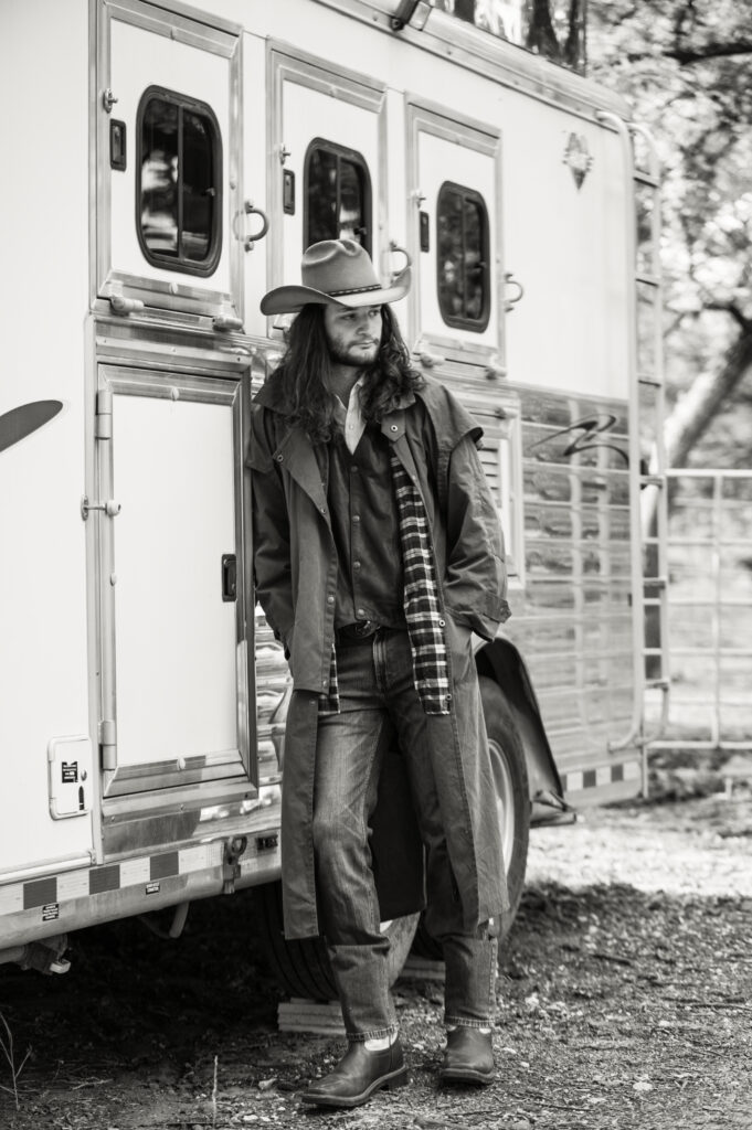 Black‑and‑white portrait of a cowboy leaning against a horse trailer at an Eastern Washington rodeo, wearing a wide‑brimmed hat, long coat, jeans, and boots — captured with a timeless, documentary‑style approach by a Washington family and headshot photographer.