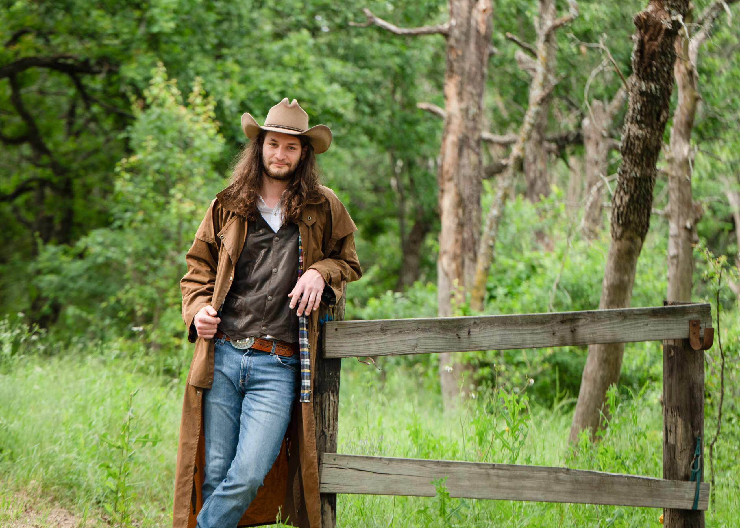 Cowboy in a long coat and hat leaning against a wooden fence in a wooded Texas ranch setting, photographed in a rustic, documentary style by an Eastern Washington family and headshot photographer.
